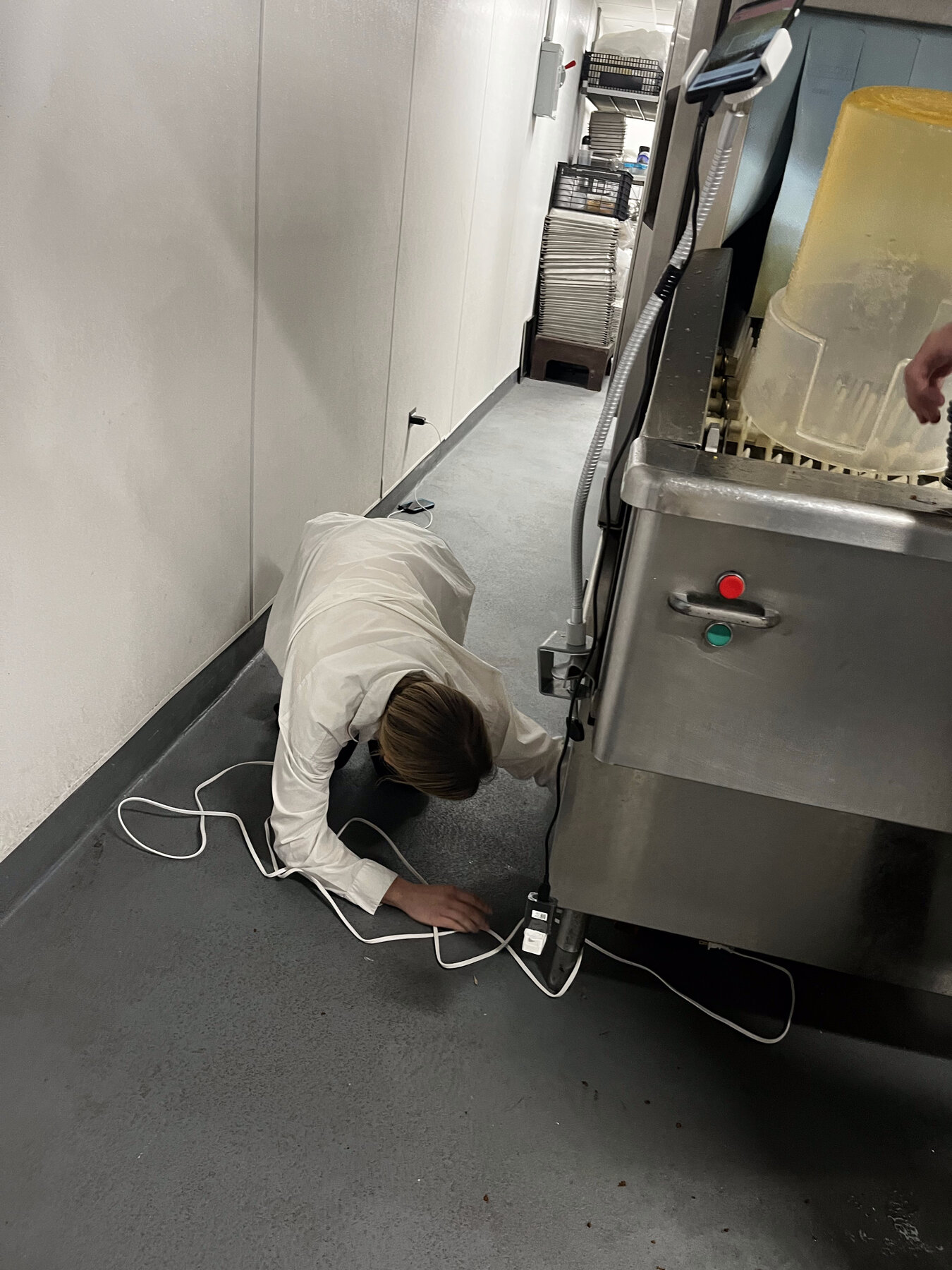 Hayley on the floor of a dishwashing room, wiring a mounted Android scanning device to a power outlet near the dish machine