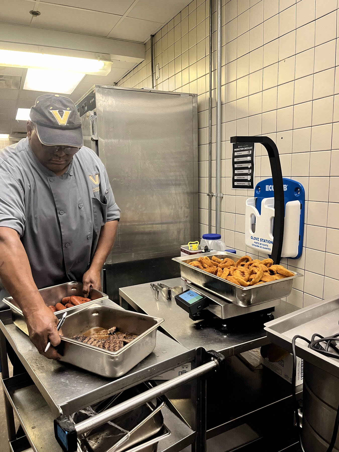 An operator in a university dining kitchen weighing a pan of onion rings on the StreamLine scale, with additional food pans staged on a cart nearby